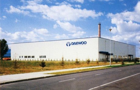 Industriehalle von Daewoo mit Firmenlogo, Straße im Vordergrund, Schornstein im Hintergrund, bewölkter Himmel.