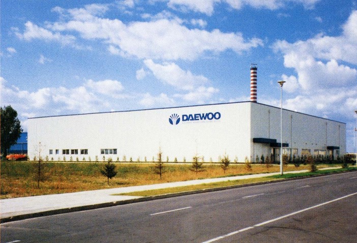 Industriehalle von Daewoo mit Firmenlogo, Straße im Vordergrund, Schornstein im Hintergrund, bewölkter Himmel.