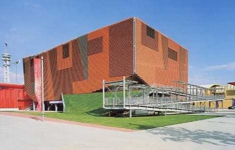 Modern building with a red façade and asymmetrical shape, surrounded by a green hill and a temporary grandstand.