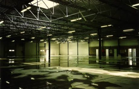 Empty industrial hall with artificial lighting, reflective concrete floor and roof construction made of steel beams.