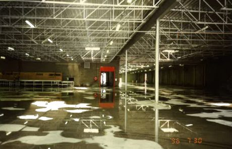 Interior view of an empty industrial hall with metal beams and reflective puddles on the concrete floor.
