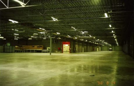 Interior view of an empty industrial hall with concrete floor, ceiling lighting and red door at the end.
