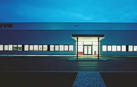 A modern industrial building at dusk with a central entrance door and illuminated company lettering above the door.