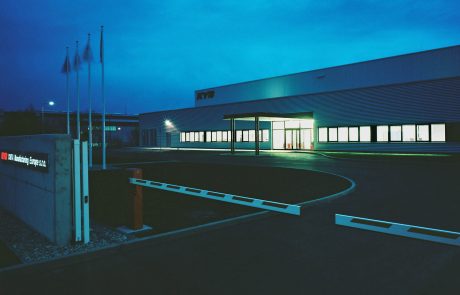 Industrial building at dusk with an illuminated entrance area and curved access, surrounded by flag masts.
