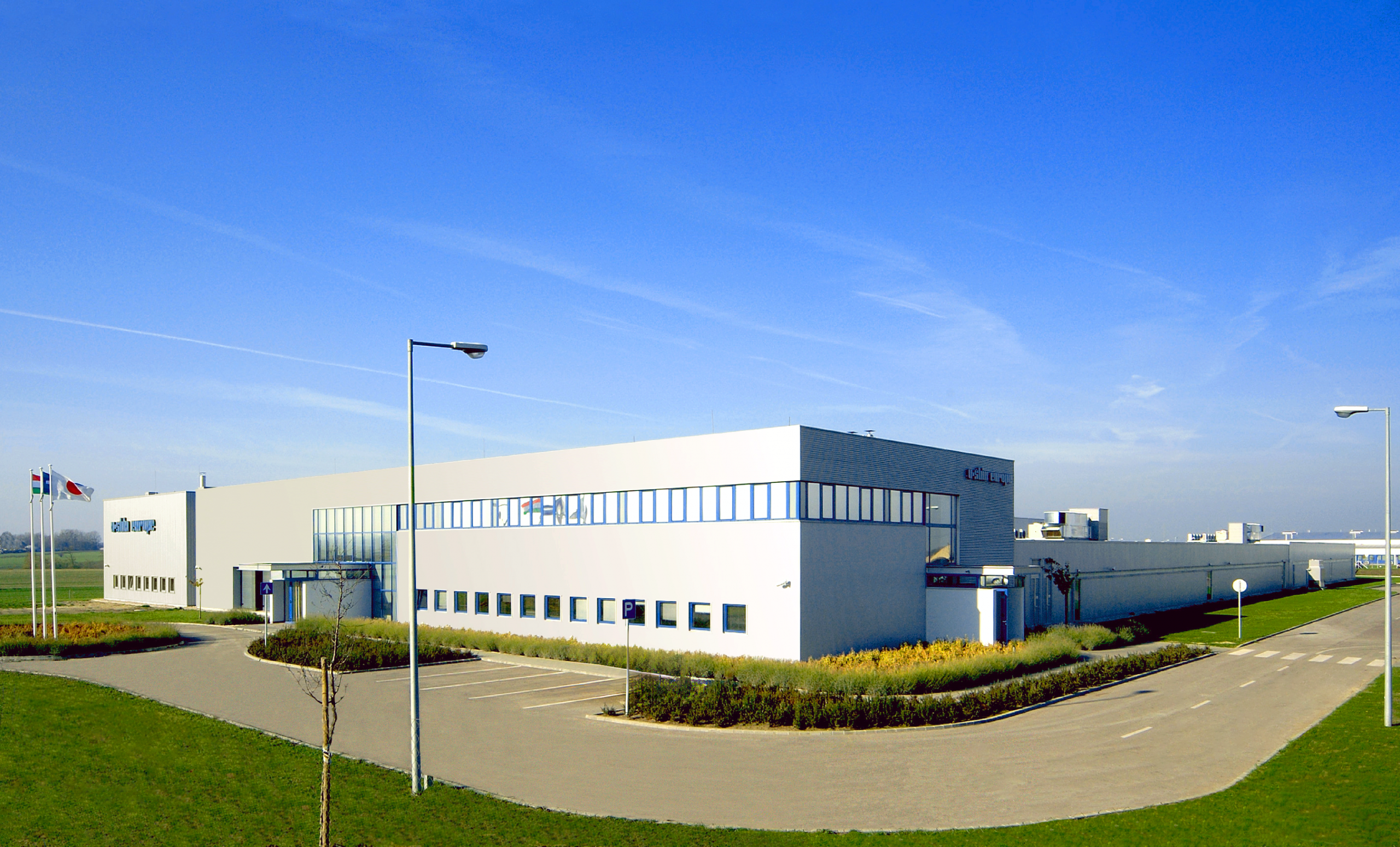 Modern commercial complex with flat roofs, consisting of several connected white buildings with extensive window fronts and striking blue accents. The complex is surrounded by manicured lawns and a paved path leading to a main entrance. Flagpoles with hoisted flags are positioned in front of the building. U-Shin Factory built by Takenaka Europe in 2004.