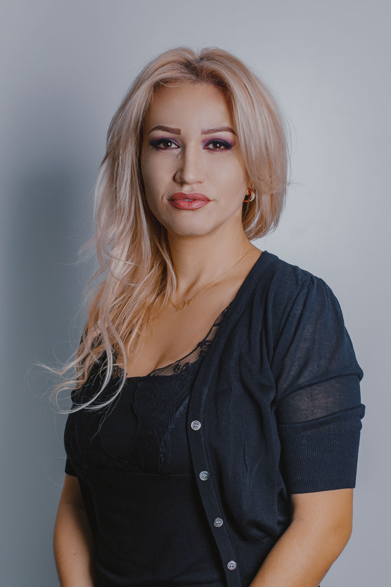 Ciuca Izabela Woman with blond hair, black clothes and smile stands in front of a gray background.