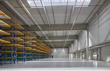 Leere Industriehalle mit gelben und blauen Regalen links, glänzendem Boden und Decke mit Leuchtstofflampen.