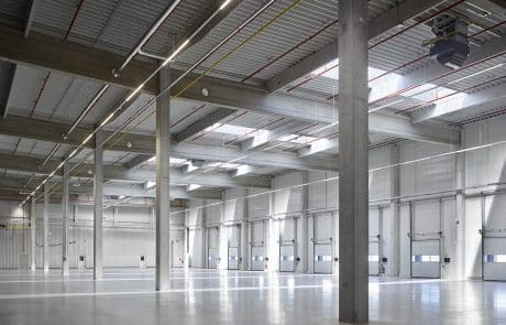 Große, leere Industriehalle mit grauem Boden, weißen Wänden und Säulen, Beleuchtung an der Decke.