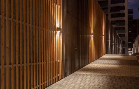 Illuminated walkway next to a wall with a vertical wooden slat structure at dusk.