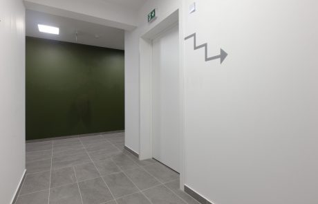 Modern office hallway interior with smooth white walls, gray floor tiles and a green accent wall. Lighting comes from wall lights and ceiling spotlights, with an arrow symbol on the right in the picture.