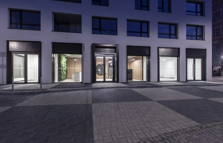 Building facade at night, with symmetrical white window frames, glass entrance doors and subtle lighting highlighting the building and cobblestone walkways.