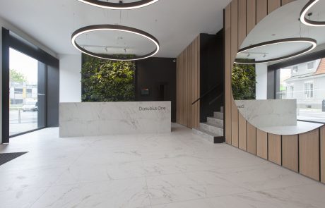 Modern designed entrance hall of an office building with white marble floor, wooden panels on the walls and futuristic circular ceiling lights. One of the reception desks reads “Danubius One.”