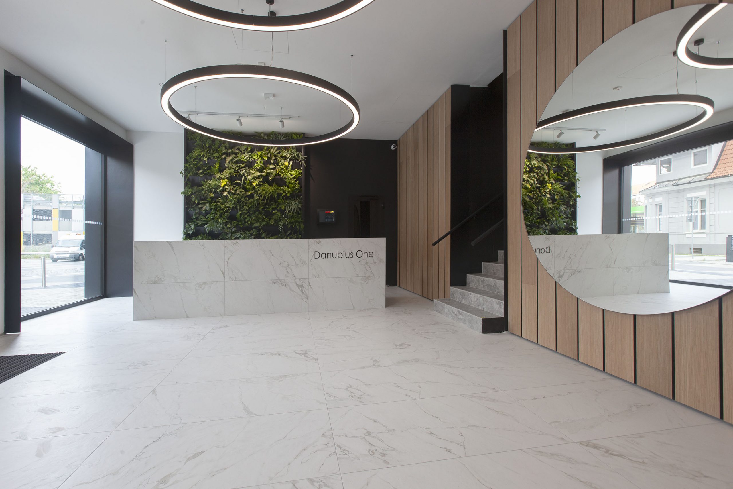 Modern designed entrance hall of an office building with white marble floor, wooden panels on the walls and futuristic circular ceiling lights. One of the reception desks reads “Danubius One.”