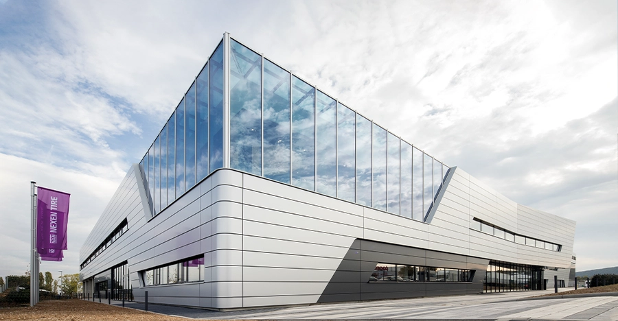 Modernes Gebäude mit Glasspitze, spiegelnden Fenstern und klarer Linienführung unter bewölktem Himmel.