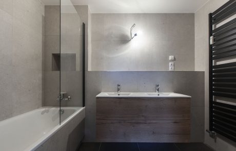 Modern bathroom with a combination of light tiled walls, a dark wooden unit with two sinks and a bathtub. Radiator on the right wall.