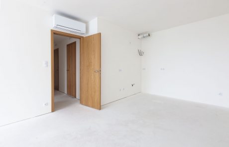 Bright, minimalist interior design of an unfinished room with a wooden door frame in the middle and concrete-like flooring. Right wall offers several sockets, ceiling with installed air conditioning unit.