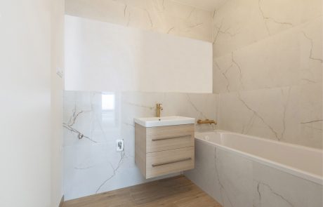 Modern bathroom with light marble tiles, white walls and minimalist design. Features include a bathtub, a wall-mounted bathroom sink with a light wood base cabinet and a gold fitting.