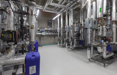 Modernly equipped machine room with numerous metal pipes, valves and insulation, illuminated by ceiling spotlights, concrete floor.