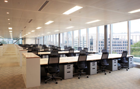 Modern office with numerous desks, bright ceiling lights and floor -to -ceiling windows with city views.