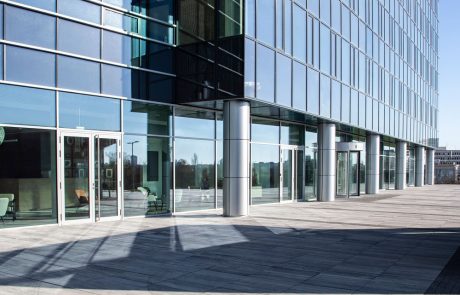 Moderne Glasfassade eines Bürogebäudes mit reflektierenden Fenstern und metallischen Säulen bei Tageslicht.