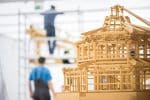 Wooden model of a traditional building in the foreground, blurred depiction of workers in the background.