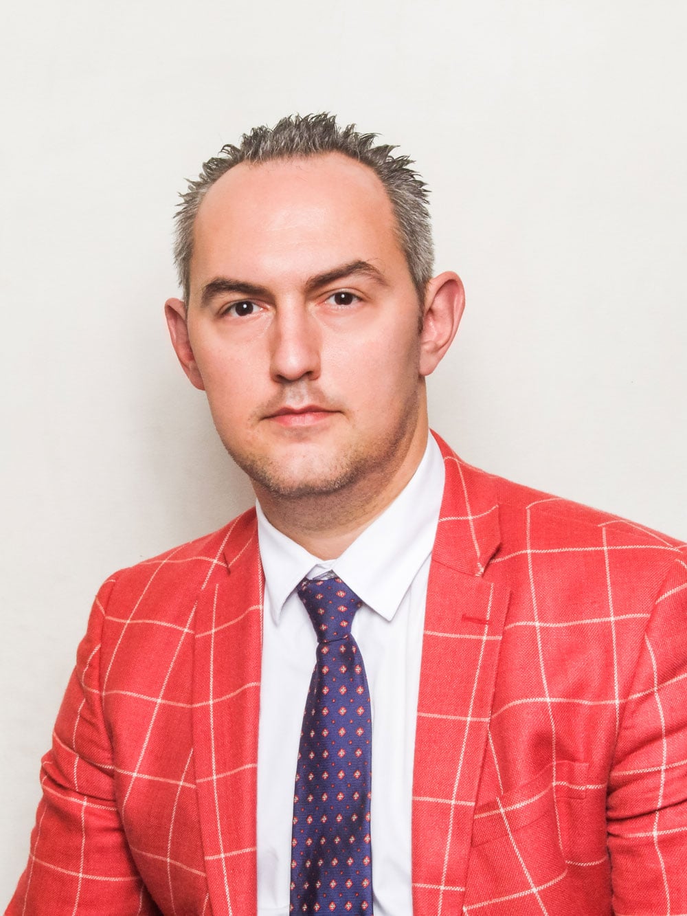 Portrait of a man Mihajlovic Uros with short hair in a red checked jacket, white shirt and patterned tie against a light background.