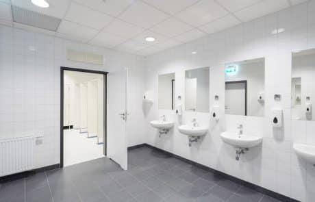 Moderne, saubere Toilettenanlage mit mehreren Waschbecken, Spiegeln und Trennwänden; neutrale Farbtöne und hell beleuchtet.