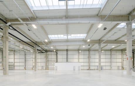 Empty industrial hall with white walls, concrete columns and bright lighting through ceiling windows.