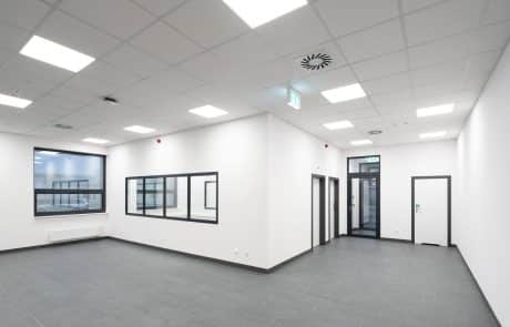 Interior of a modern office with white walls, gray tiles, recessed lights in the ceiling panel and windows to the side.