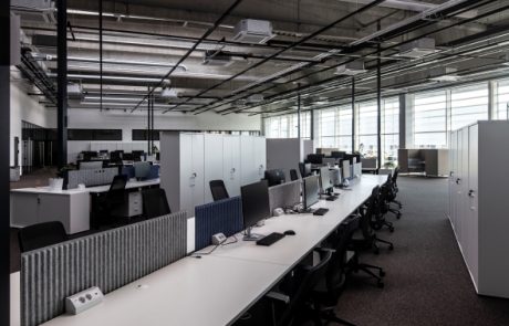 Modern office landscape with open space design, various workstations with monitors and partitions. Ceiling structure with visible cables and lighting devices, windowed exterior walls.
