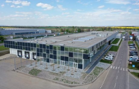 Aerial view of a modern production hall with a glass facade, equipped with solar panels on the flat roof, surrounded by parked cars and planted green areas on a street.