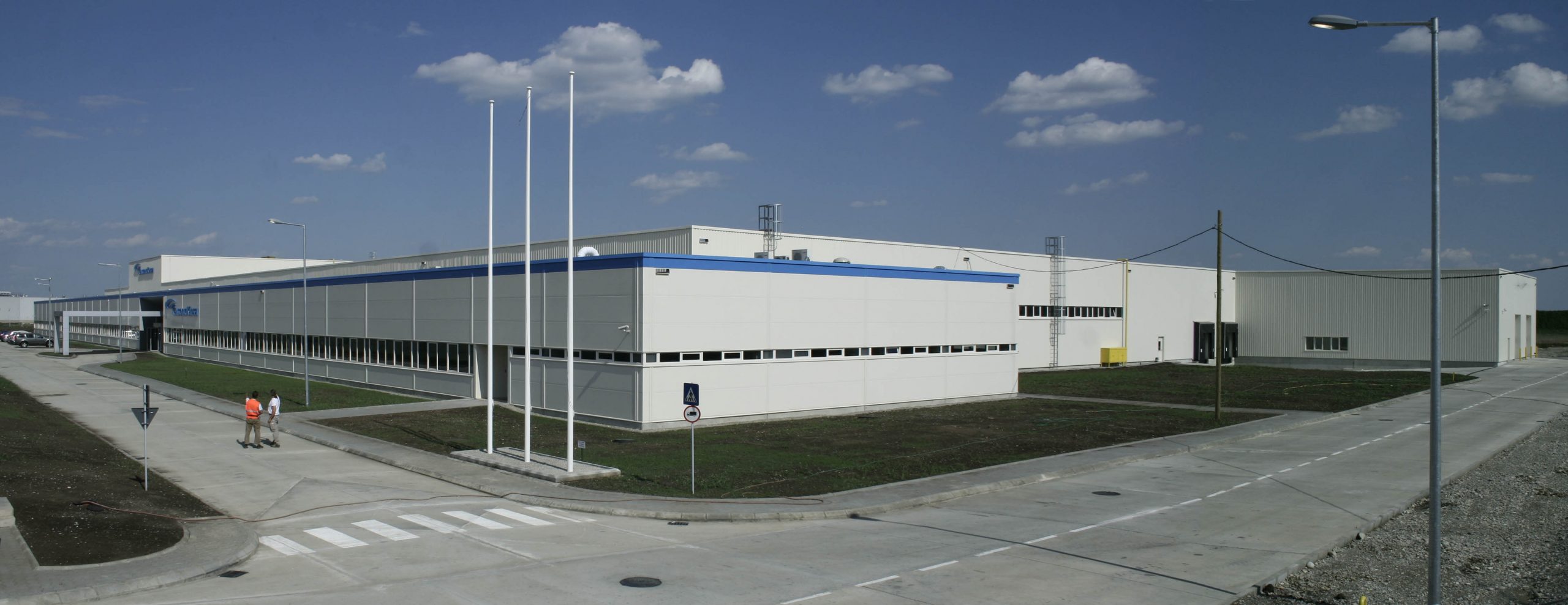 Industriekomplex mit modernem Aussehen, weiße und blaue Fassade, unter blauem Himmel, Personen im Vordergrund.