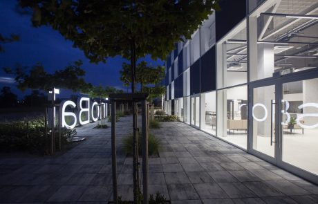 Nighttime view of a modern office building with large glass fronts and an illuminated forecourt. Trees line the illuminated walkway made of large-format tiles.