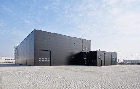 Modern industrial hall with a gray façade under a blue sky, surrounded by a large, paved forecourt.
