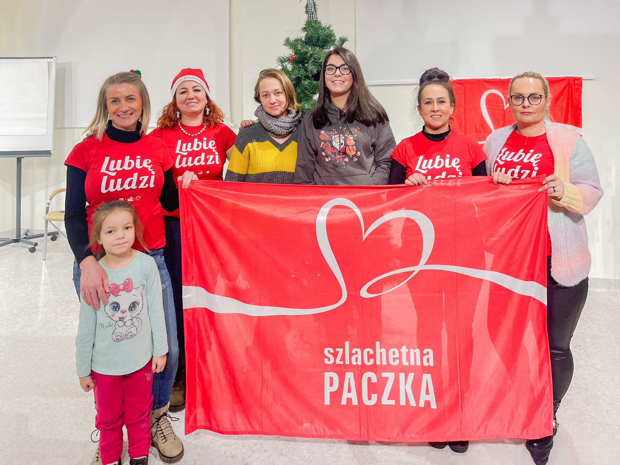 Grupa wolontariuszy w czerwonych koszulkach trzymających baner akcji "Szlachetna Paczka" obok choinki.