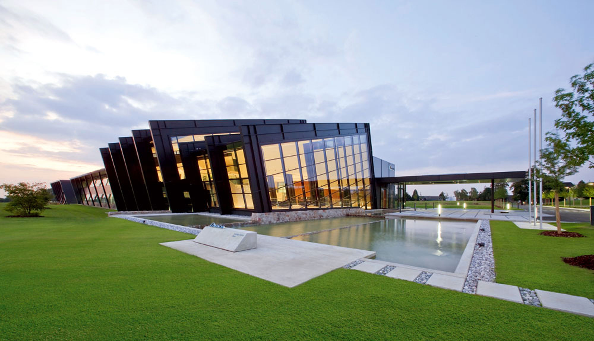 Modern office building complex with large glass façade, surrounded by manicured lawns and a water basin at dusk.