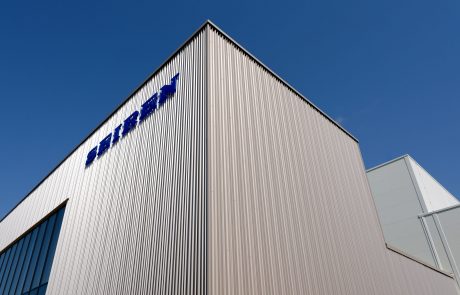 View of a modern industrial building with vertical façade cladding and blue company lettering against a clear sky.