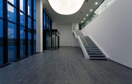 Modern office lobby with shiny floor tiles, white staircase, dark window frames and artistic ceiling lighting.