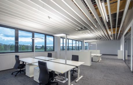 Modernes Büro mit weißen Tischen, schwarzen Stühlen, großen Fenstern und heller Beleuchtung.