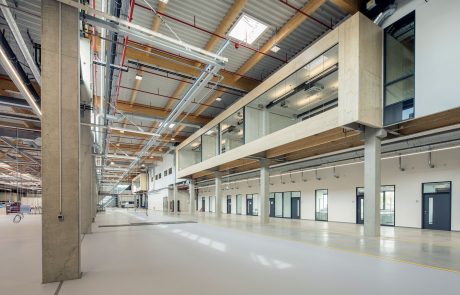 Moderne Industriehalle mit sichtbaren Betonsäulen, Holzbalken, Installationen an der Decke und großen Fenstern.