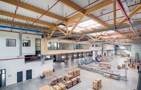 Große Industriehalle mit hohen Decken, Holzträgern, Oberlichtern und verpackten Waren auf dem Boden.