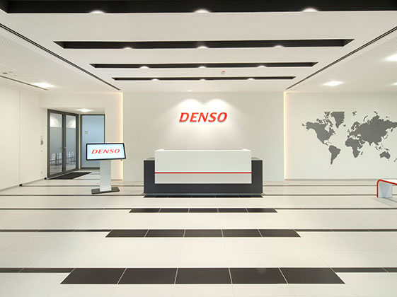 Empfangsbereich mit dem Logo von DENSO an der Wand, einer Weltkarte, beleuchtetem Tresen und modernem Bodendesign.