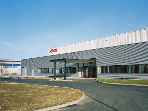 Moderne Industriehalle von KYB mit Firmenlogo, großer Eingangstür und gepflegter Außenanlage bei Tageslicht.