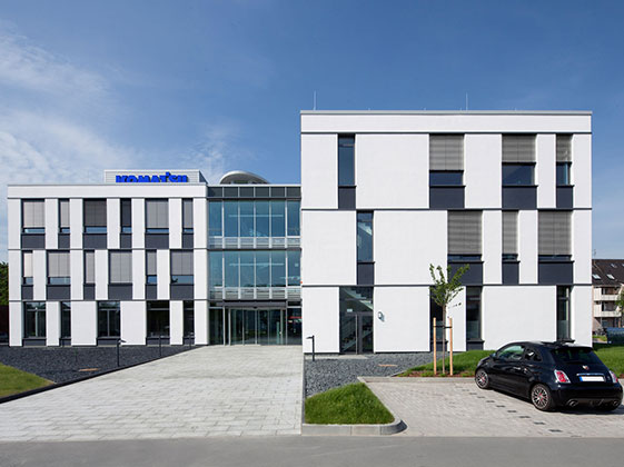 Modernes Bürogebäude mit weißer Fassade, großen Fenstern und Firmenlogo unter blauem Himmel.
