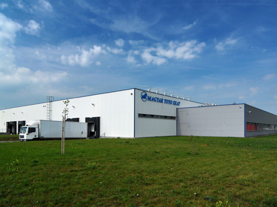 Industriegebäude der Magyar Toyo Seat Fabrikerweiterung bei Tageslicht mit blauem Himmel und Grünfläche im Vordergrund.